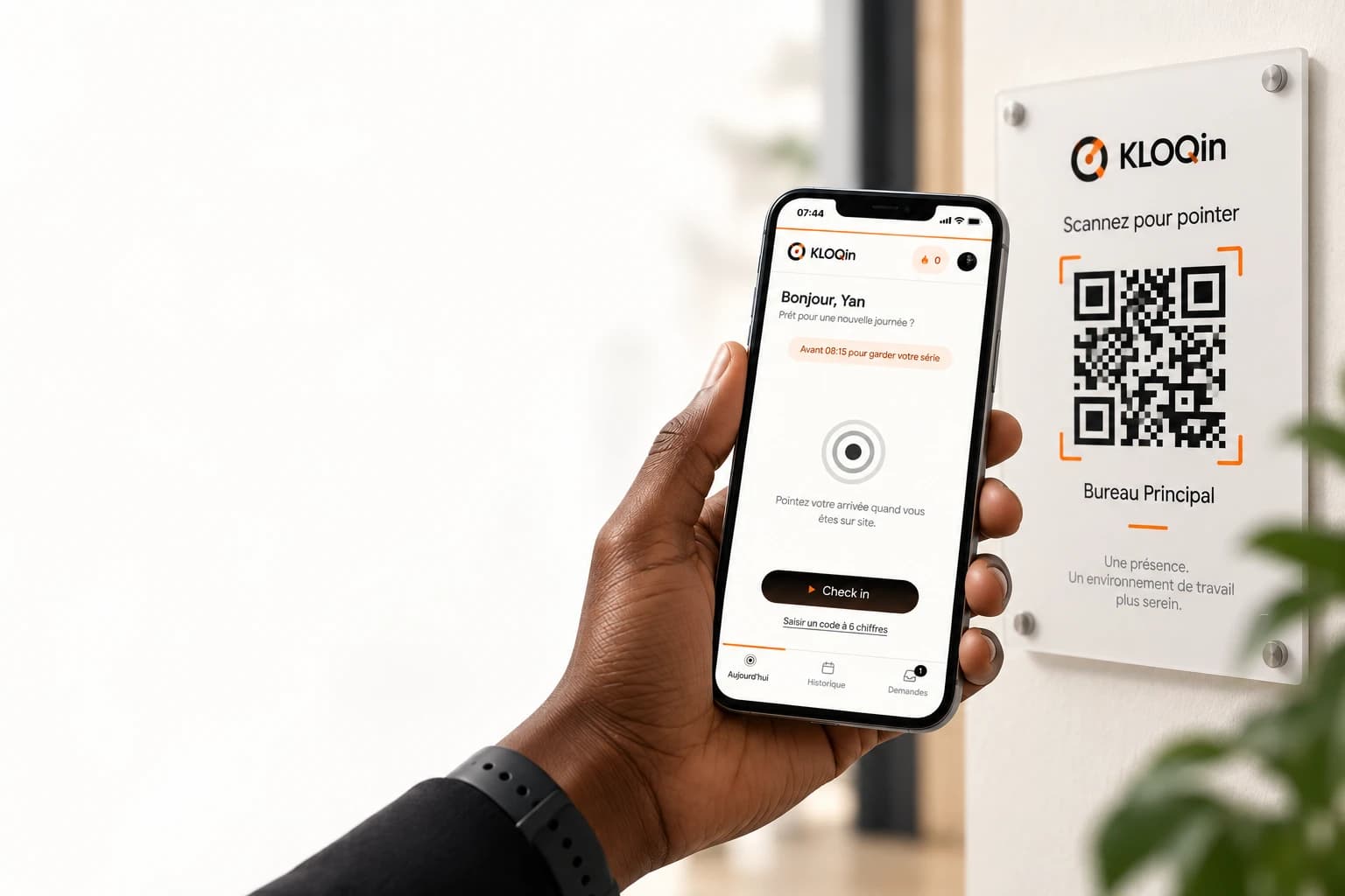 Une main tient un téléphone affichant l’écran d’arrivée de KLOQin, devant une plaque murale « Scannez pour pointer » portant un QR code et le nom du site, Bureau Principal.